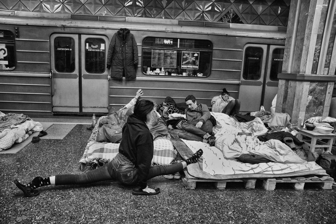 Schuilende burgers op matrassen in een metrotunnel; vrouw doet spagaat naast trein en geïmproviseerde bedden.