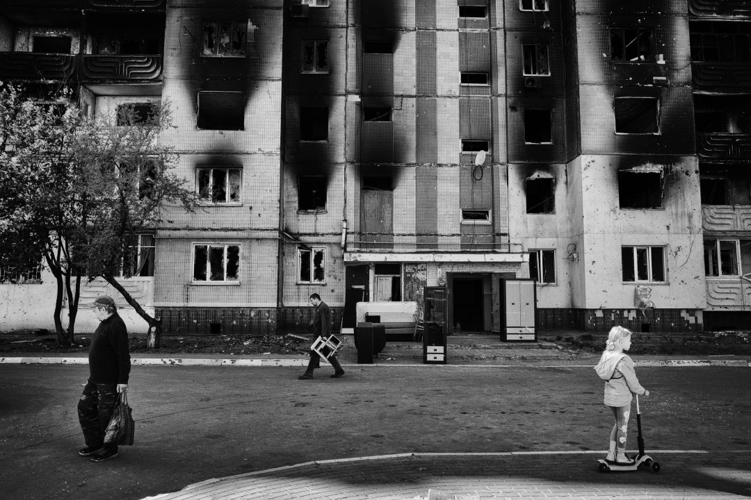 Verwoest flatgebouw met zwartgeblakerde ramen; mannen lopen langs, kind op step kijkt naar de ravage